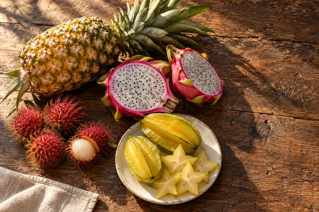 Abacaxi, pitaya aberta, rambutãs e carambolas em prato claro sobre mesa de madeira iluminada pelo sol.