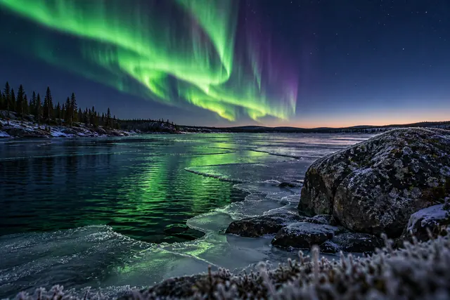 Aurora boreal verde e violeta sobre lago parcialmente congelado, com rochas cobertas de geada e coníferas na margem.