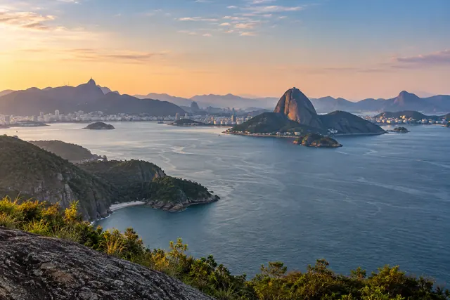 Vista da Baía de Guanabara ao amanhecer, com Pão de Açúcar, montanhas, águas azuis e vegetação em primeiro plano.