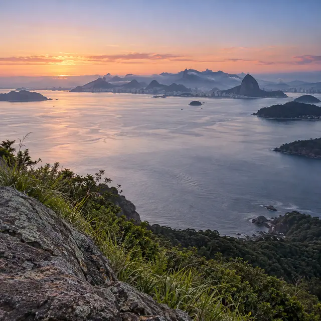 Baía de Guanabara ao amanhecer, com pedras e mata em primeiro plano, ilhas e montanhas sob névoa ao fundo.