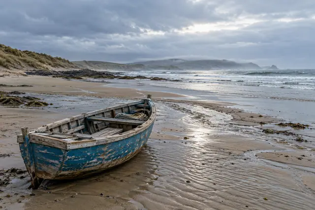 Barco de pesca azul na areia molhada, com ondas, rochas e dunas ao fundo sob um céu nublado.