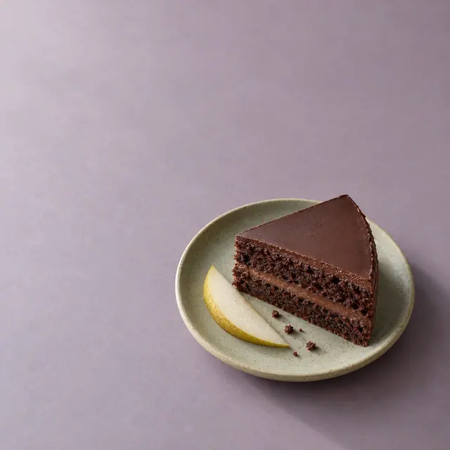Fatia de bolo de chocolate e lasca de pera em prato verde-claro sobre fundo lavanda, com amplo espaço livre.