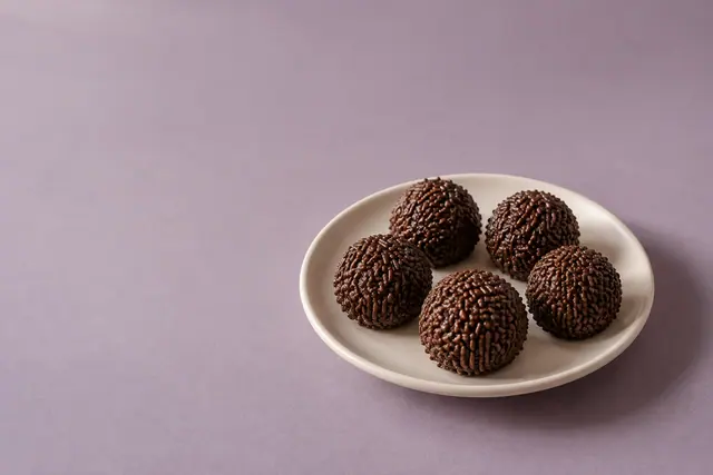 Cinco brigadeiros com granulado em prato claro sobre fundo lavanda, com luz suave e amplo espaço livre à esquerda.