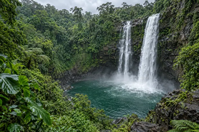 Cachoeira em duas quedas sobre paredão escuro, com poço verde e vegetação tropical densa ao redor.