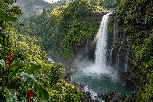 Cachoeira em paredão rochoso cercado por floresta tropical, com água verde-azulada e helicônias em primeiro plano.
