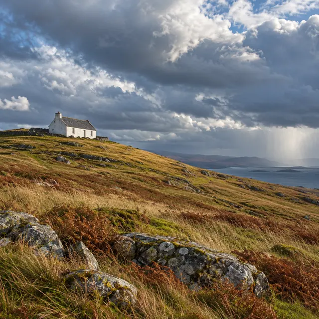 Casa branca no alto de colina com capim dourado e pedras, sob nuvens densas e chuva sobre o mar ao fundo.