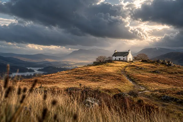 Casa branca em colina dourada sob nuvens escuras e raios de sol, com trilha, lago e montanhas ao fundo.