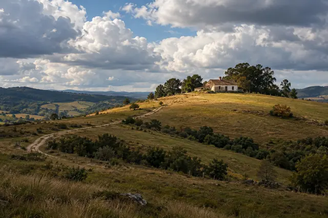 Casa rural no alto de uma colina com pastagens douradas, estrada sinuosa e grandes nuvens no céu.