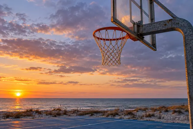 Cesta de basquete em quadra azul à beira-mar, com dunas e céu alaranjado e lilás ao pôr do sol.