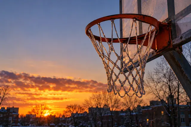 Cesta de basquete vista de baixo ao pôr do sol, com árvores sem folhas e prédios desfocados ao fundo.