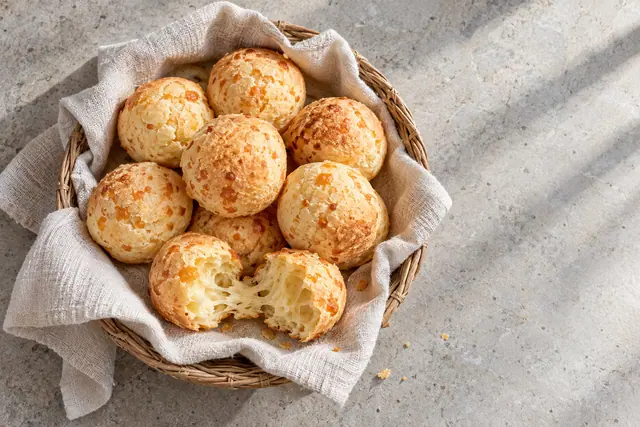 Cesta com pães de queijo dourados e um pão aberto sobre tecido claro, em bancada de pedra iluminada pelo sol.