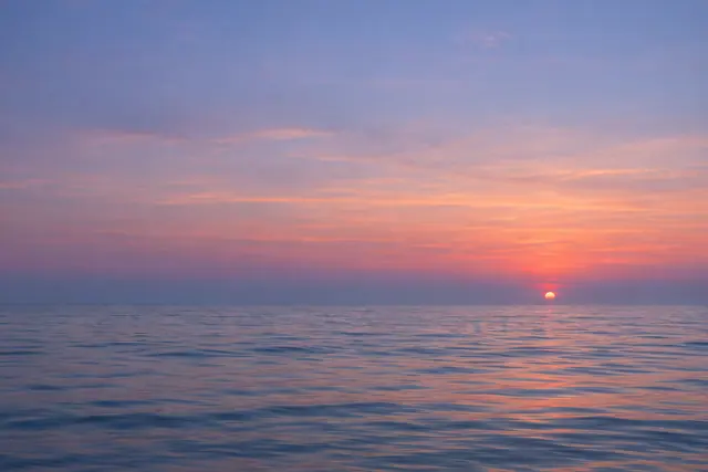 Sol próximo ao horizonte sobre mar calmo, com céu amplo em tons de rosa e lavanda e suaves reflexos na água.