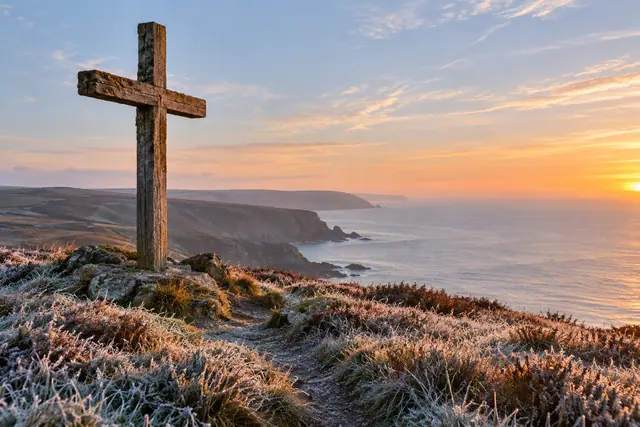 Cruz de madeira em colina com geada diante do mar e de falésias sob a luz dourada do amanhecer.