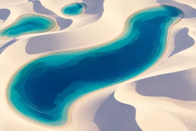 Vista aérea ilustrada de uma lagoa azul curva e duas lagoas menores entre dunas claras com sombras lilases.