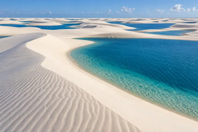 Dunas brancas com marcas do vento cercam lagoas azuis e turquesa sob céu claro nos Lençóis Maranhenses.