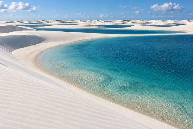Dunas brancas cercam lagoas azuis e transparentes sob céu com nuvens esparsas, em paisagem inspirada nos Lençóis Maranhenses.