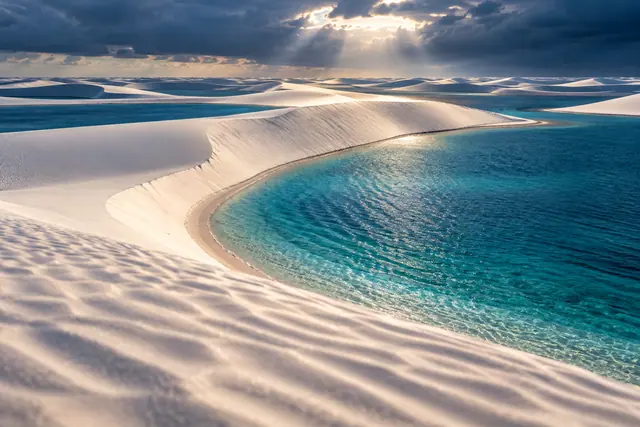 Dunas brancas contornam lagoas azuis sob nuvens escuras e raios de sol, em paisagem inspirada nos Lençóis Maranhenses.