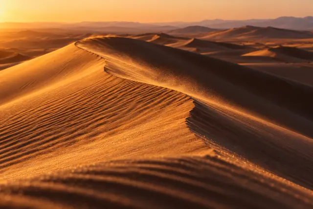 Dunas de areia dourada ao entardecer, com cristas sinuosas, sombras profundas e grãos levados pelo vento.