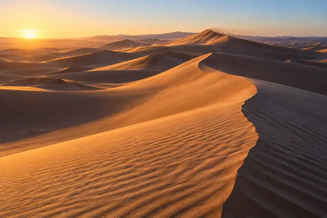 Dunas de areia com cristas douradas, ondulações e sombras longas sob o sol baixo no horizonte.