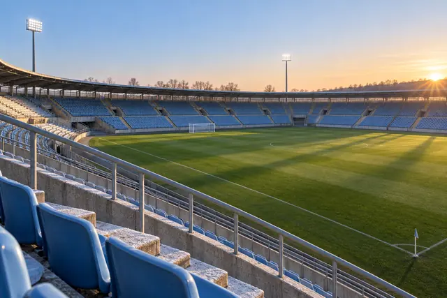 Estádio vazio ao amanhecer, com cadeiras azuis, refletores acesos e sombras longas sobre o gramado.