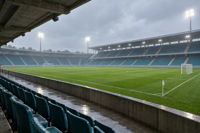 Estádio de futebol vazio sob chuva, com refletores acesos, gramado verde e assentos azulados molhados.