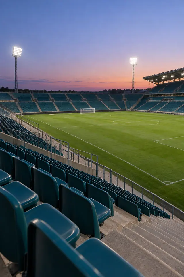 Estádio de futebol vazio com assentos azul-petróleo, gramado verde e refletores acesos ao entardecer.