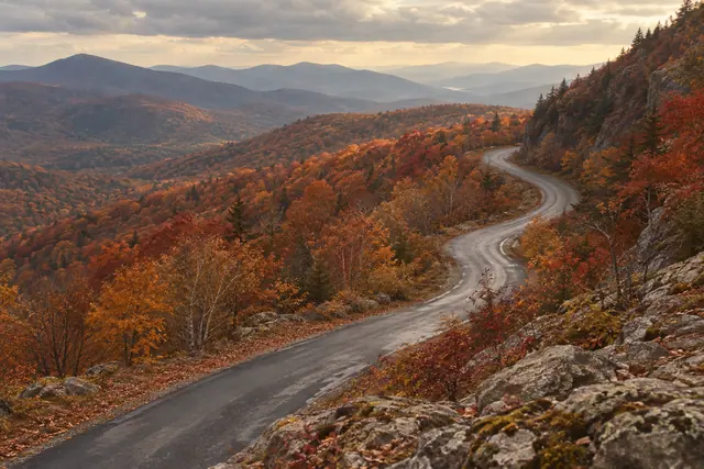Estrada sinuosa entre florestas de outono, rochas e montanhas distantes sob céu nublado em tons quentes.