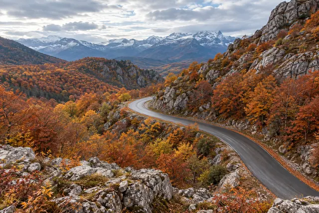 Estrada sinuosa entre rochas e árvores de outono, com montanhas nevadas ao fundo sob céu nublado.