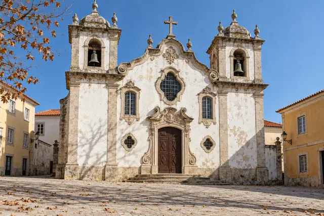 Igreja barroca com duas torres e porta de madeira diante de praça com folhas secas, sob céu azul.