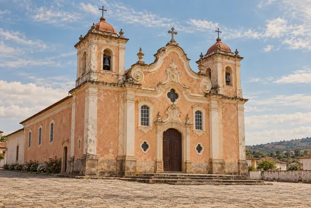 Igreja histórica com fachada pêssego, duas torres e porta de madeira diante de um largo de pedra sob céu azul.