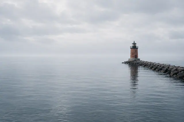 Farol de tijolos na ponta de um quebra-mar de pedras, com reflexo no mar calmo sob céu cinzento.