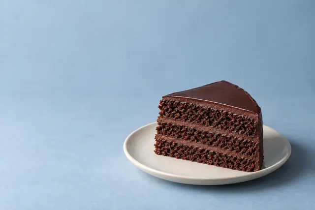 Fatia de bolo de chocolate com recheio e cobertura em prato claro sobre fundo azul, com amplo espaço vazio à esquerda.
