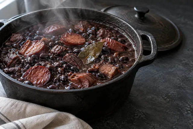 Feijoada fumegante com feijão-preto, carnes, linguiça e louro em panela de ferro sobre bancada escura.