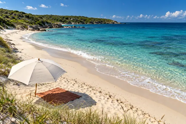 Guarda-sol claro e toalha terracota na areia de uma praia com mar turquesa e vegetação costeira ao fundo.