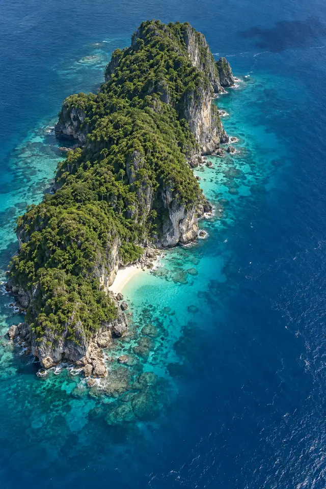 Vista aérea de ilha com mata verde, falésias e pequena praia cercada por águas turquesa e mar azul profundo.