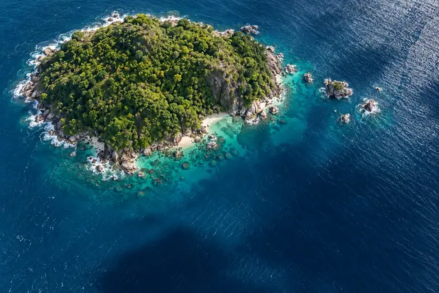 Vista aérea de ilha tropical com mata densa, praias pequenas e rochas cercadas por mar turquesa e azul profundo.