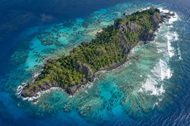 Vista aérea de ilha rochosa com vegetação verde, recifes em águas turquesa e ondas brancas ao redor.