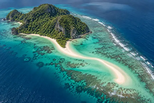 Vista aérea de ilha tropical com vegetação densa, faixa curva de areia e recifes em águas turquesa.