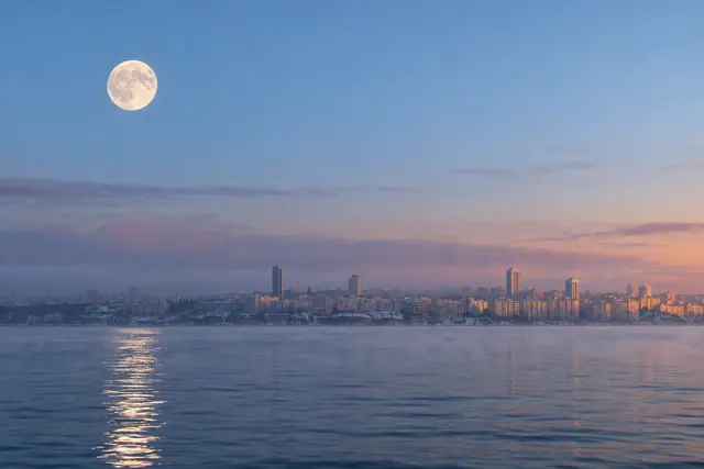Lua cheia reflete na água diante de uma cidade litorânea com névoa e prédios iluminados pelo amanhecer.