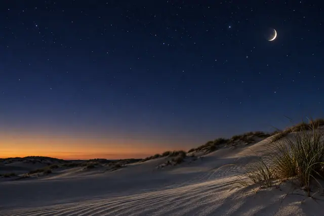 Lua crescente no céu estrelado sobre dunas de areia, com vegetação à direita e brilho alaranjado no horizonte.