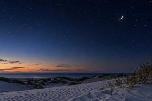 Lua crescente e estrelas sobre dunas com vegetação, mar ao fundo e uma faixa alaranjada no horizonte.