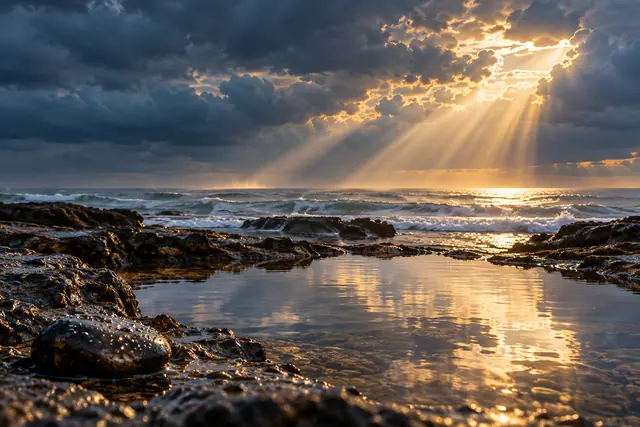 Raios dourados atravessam nuvens escuras sobre o mar e refletem em uma piscina natural entre rochas.