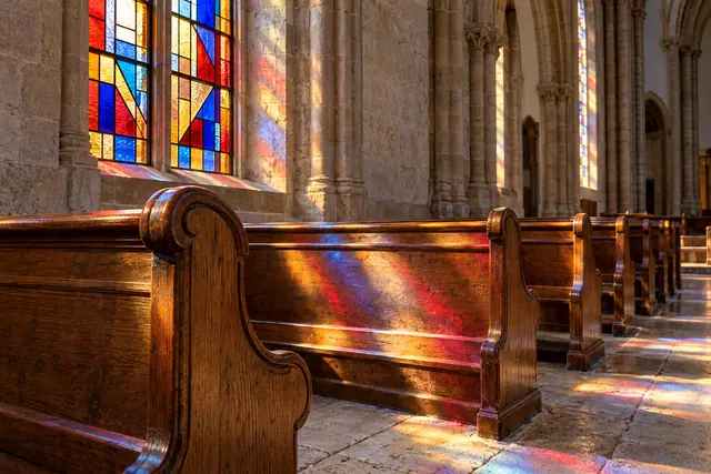 Luz de vitrais coloridos ilumina bancos de madeira e o piso de pedra de uma igreja vazia.