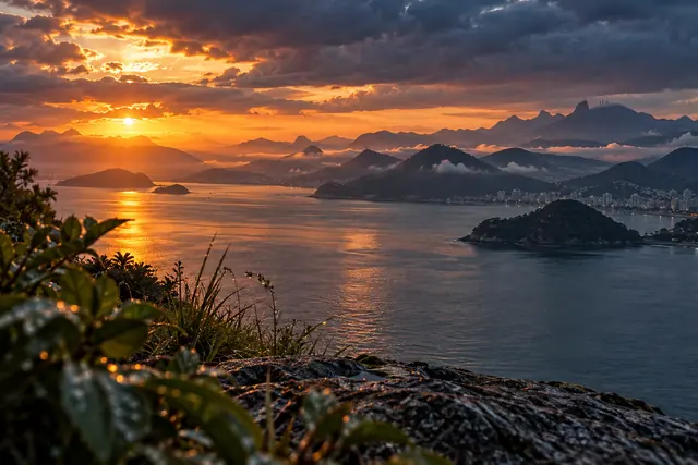 Sol entre nuvens sobre a Baía de Guanabara, com montanhas na névoa, ilhas e vegetação desfocada em primeiro plano.