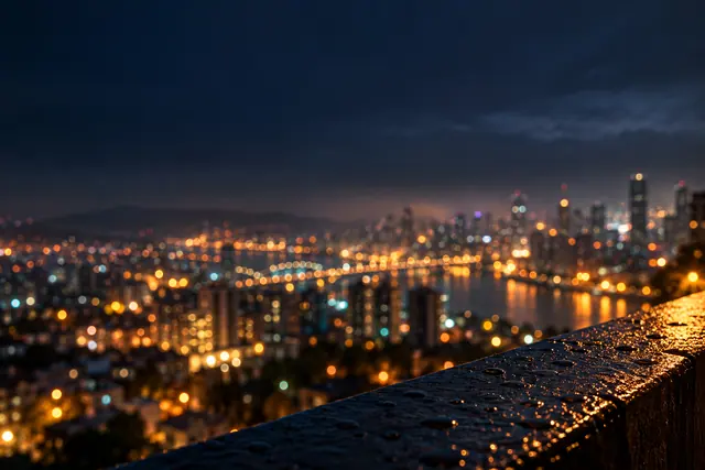 Luzes desfocadas da cidade à noite, reflexos na água e gotas de chuva sobre uma mureta sob o céu azul escuro.