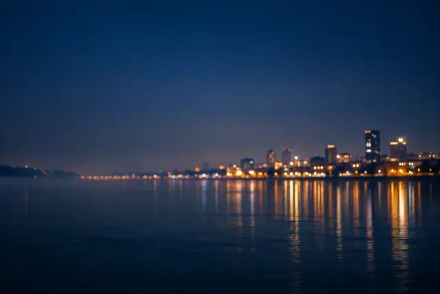 Luzes douradas da cidade em bokeh refletidas em um rio tranquilo sob um amplo céu azul-escuro.
