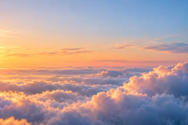 Nuvens macias iluminadas em dourado e pêssego ao amanhecer, sob um céu amplo em tons de azul.