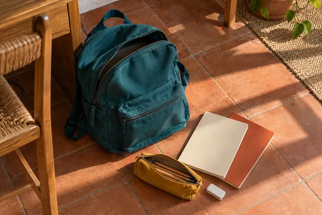 Mochila azul-petróleo, cadernos, estojo amarelo com lápis e borracha sobre piso de terracota iluminado pelo sol.