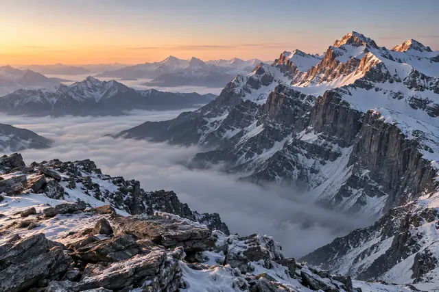 Montanhas nevadas iluminadas pelo nascer do sol, com nuvens nos vales e rochas em primeiro plano.