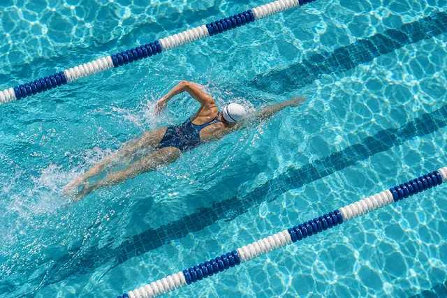 Nadadora de maiô azul e touca branca faz nado crawl em piscina com água turquesa, vista de cima.
